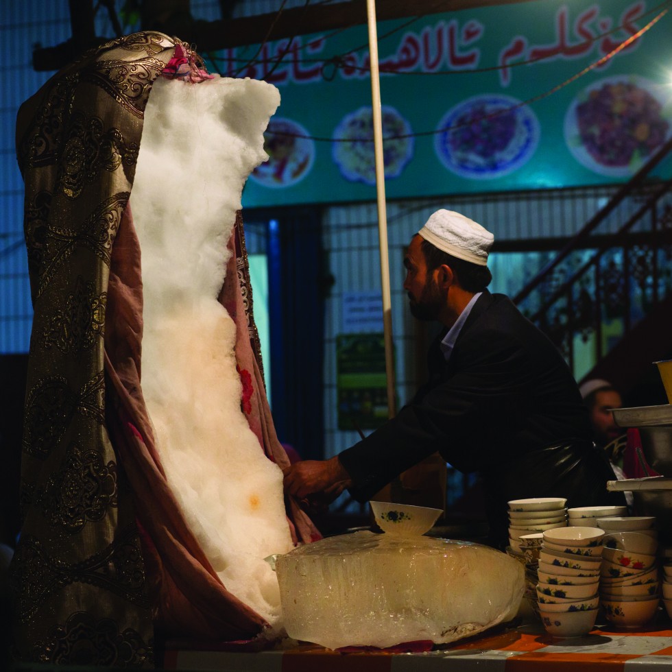 A huge piece of ice is hidden under a blanket. Starting in the middle, the vendor scrapes off some ice and tops it off with syrup.