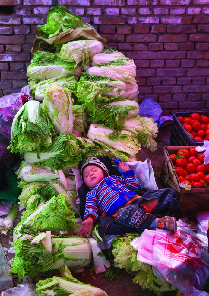 In the middle of the bustling market with dust flying and immense heat, a baby sleeps on cabbages